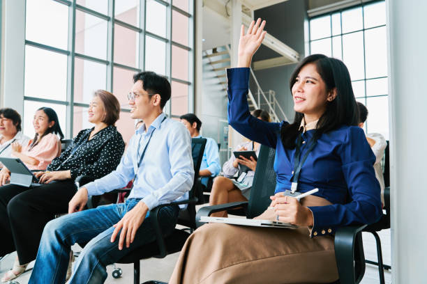 A woman in a blue blazer raises her hand actively during a seminar in a bright Convention hall at office